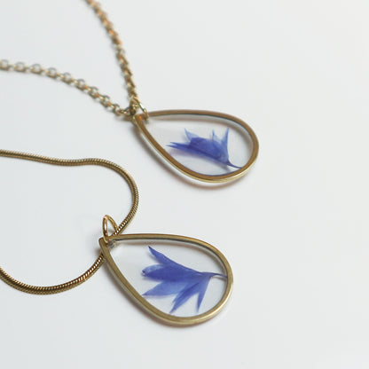 Drupt gouden druppelvormige ketting met een echte blauwe korenbloem in hars, minimalistisch en handgemaakt sieraad.