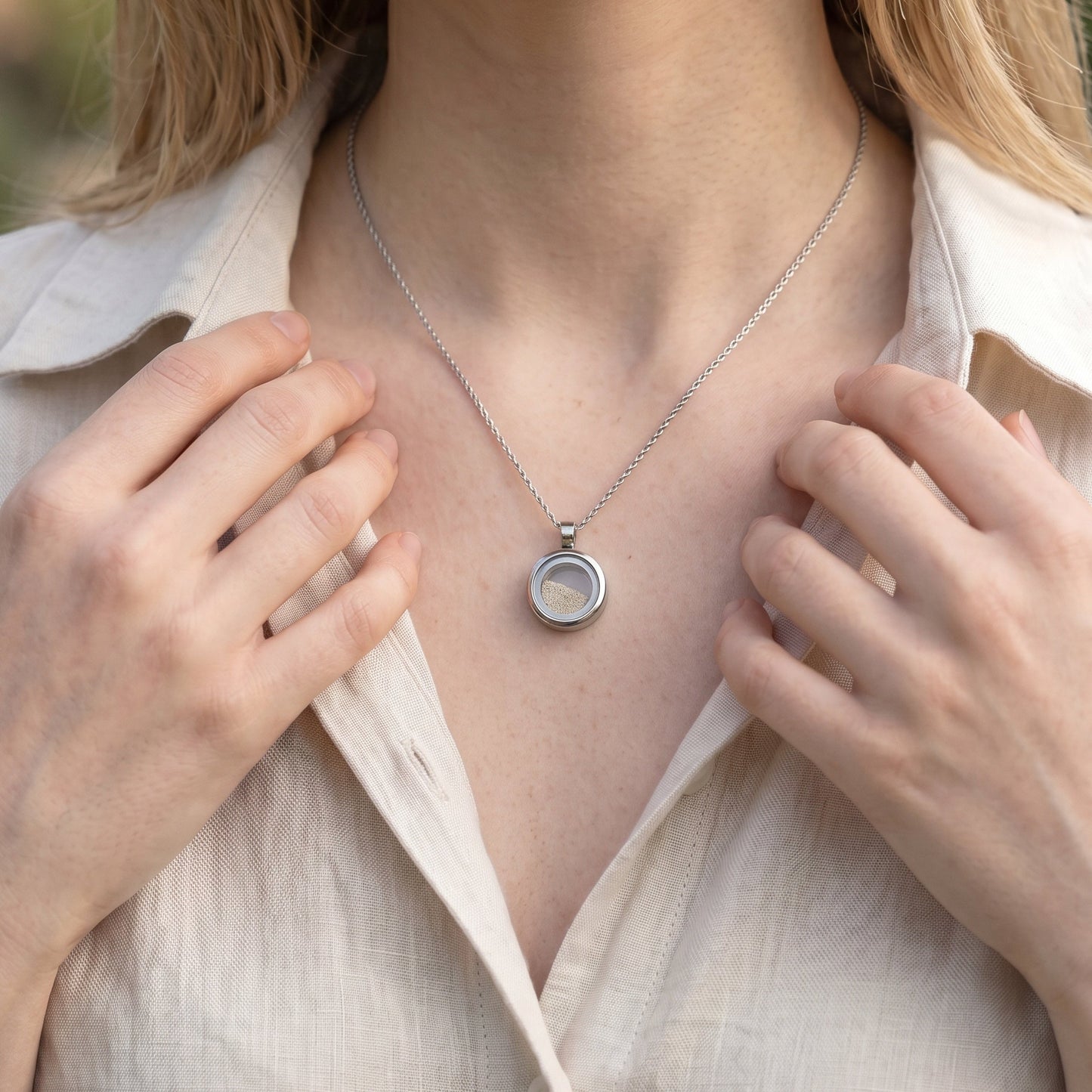 Drupt Koester zilveren gedenkhanger met zand gedragen aan een ketting door een vrouw in een beige linnen blouse.