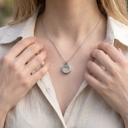Drupt Koester zilveren gedenkhanger met zand gedragen aan een ketting door een vrouw in een beige linnen blouse.
