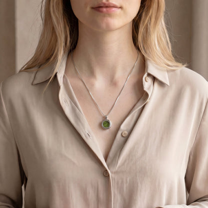 Drupt Koester zilveren hanger met groene inhoud gedragen aan een ketting door een vrouw in een beige blouse.