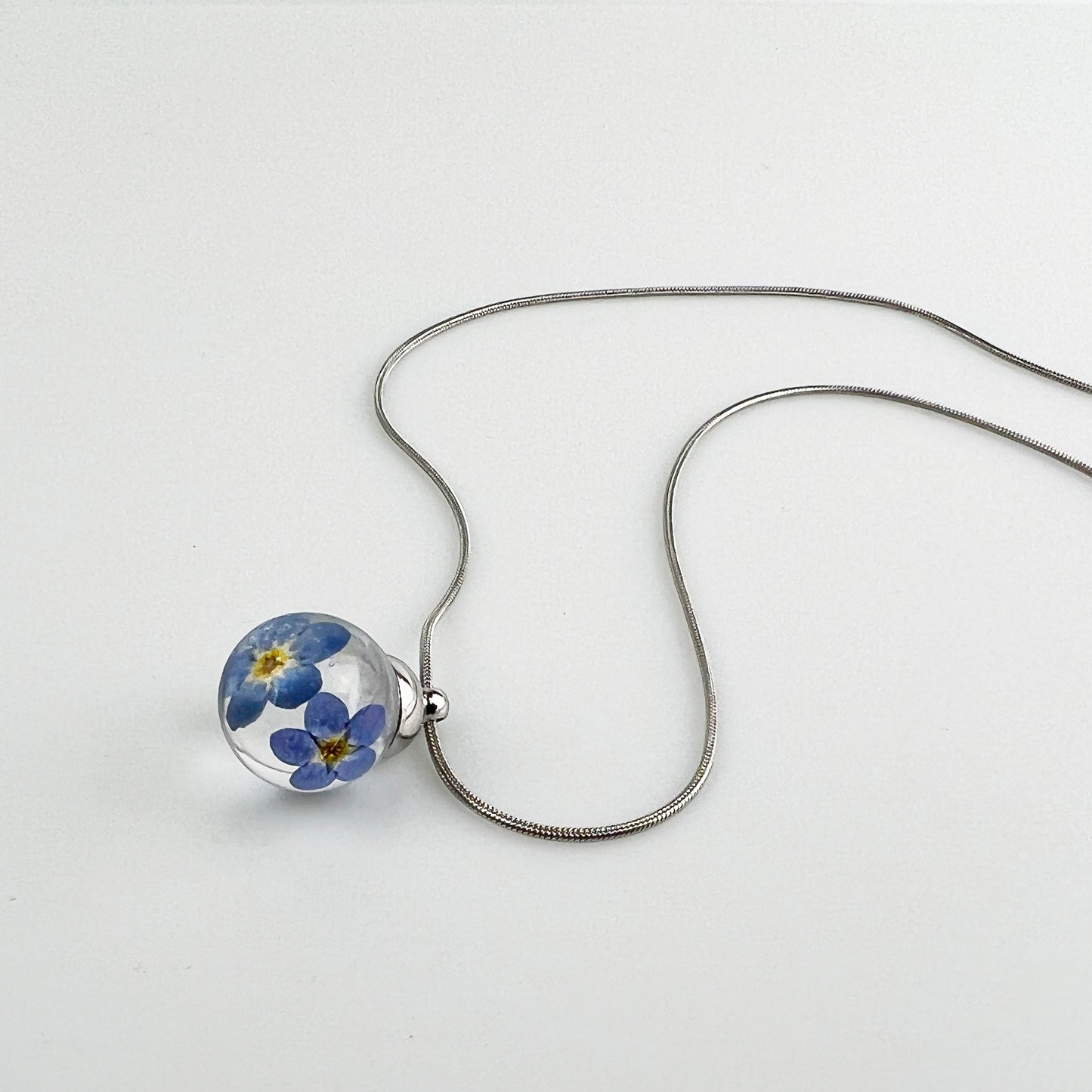 Drupt hanger met twee blauwe vergeet-mij-nietjes in een transparante bol aan een zilverkleurige ketting.