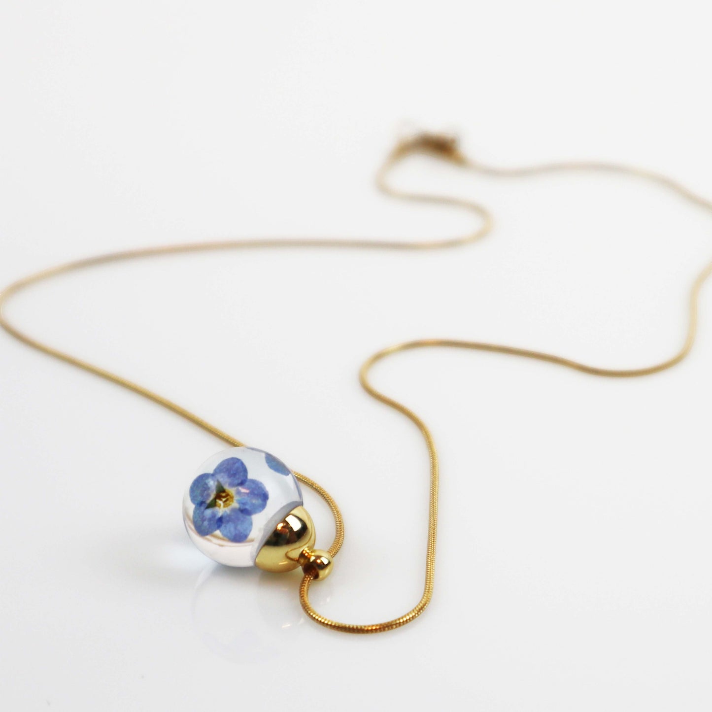 Drupt ketting met ronde glazen hanger waarin een blauw vergeet-mij-nietje is verwerkt aan een fijne gouden ketting.
