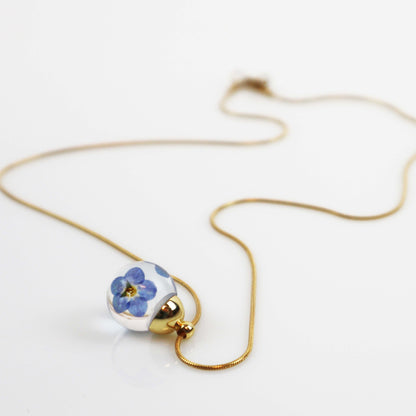 Drupt ketting met ronde glazen hanger waarin een blauw vergeet-mij-nietje is verwerkt aan een fijne gouden ketting.