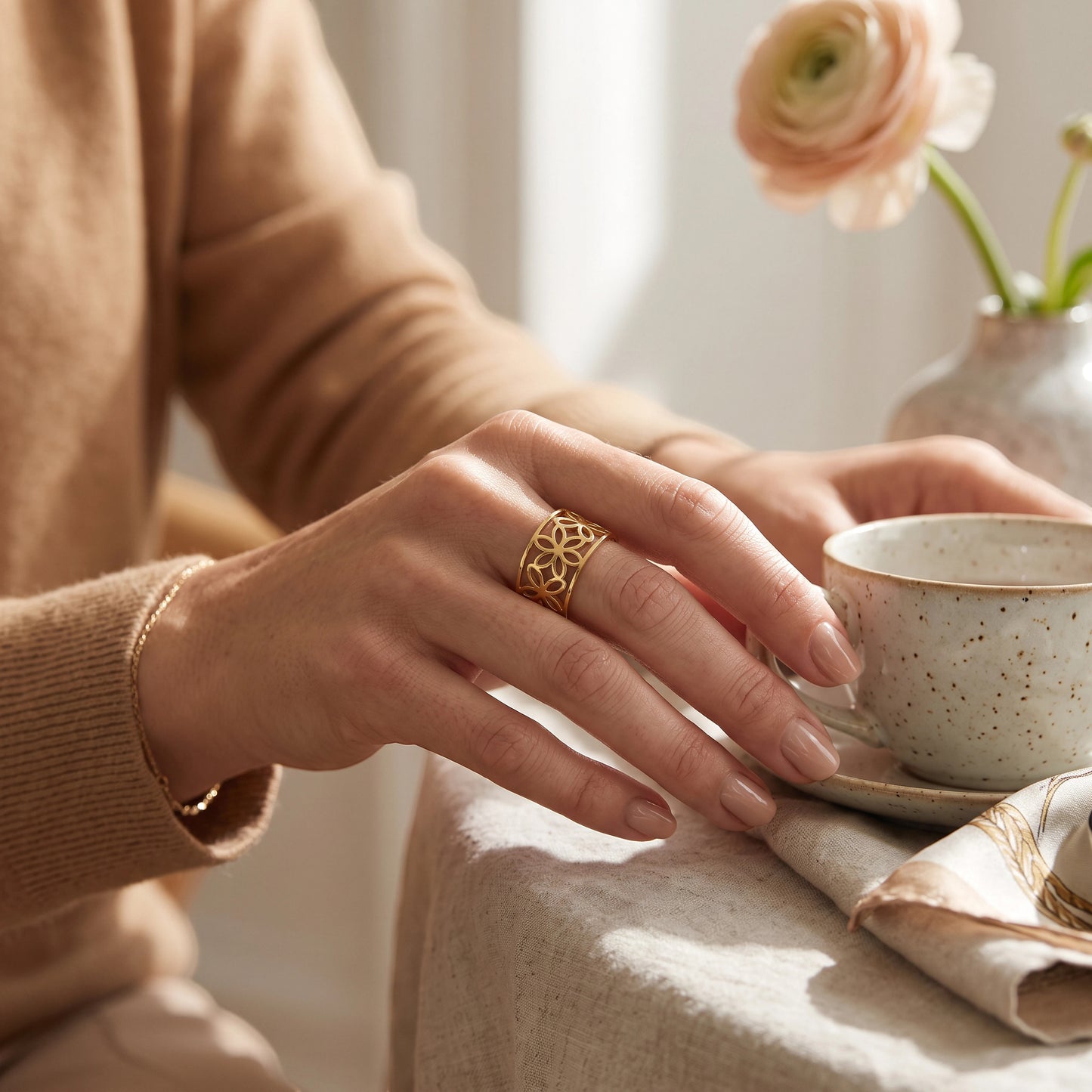 Drupt verstelbare goudkleurige ring met elegant opengewerkt bloemmotief gedragen aan een hand bij een kopje thee