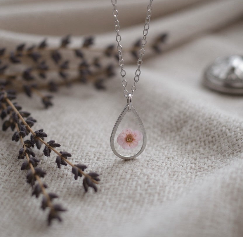 Drupt zilveren druppelvormige hanger met een echte roze pruimenbloesem aan een fijne ketting op een lichte stof.