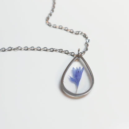 Drupt zilveren ketting met een minimalistische druppelvormige hanger en een echte blauwe korenbloem erin verwerkt.